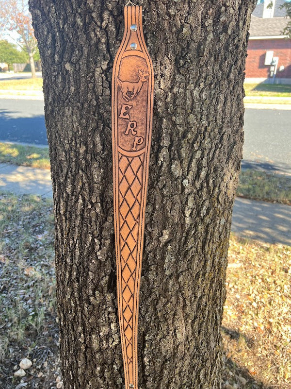 Custom hand-tooled leather gun sling with deer carving, monogram, and basket weave pattern hanging on a tree