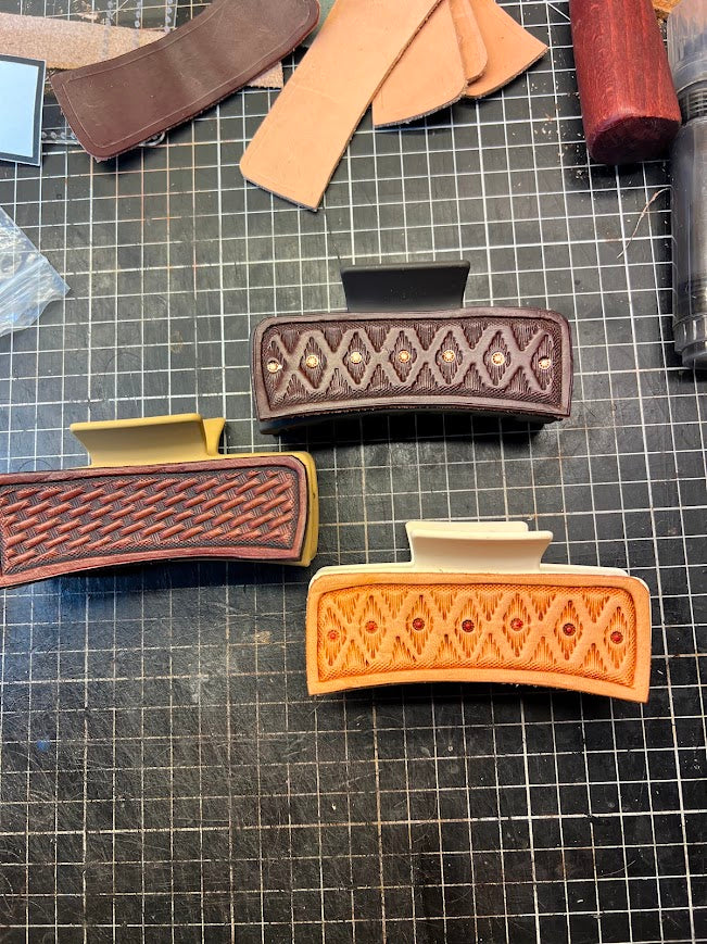 Hand-tooled leather hair clips on a workshop cutting mat showing basketweave and diamond pattern designs